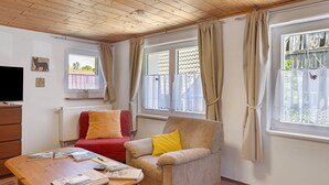 Habitació amb sostre de fusta, un sofà vermell, un butàc armau d'un color beix amb un coixí groc, una taula de fusta amb llibres i una finestra amb cortines.