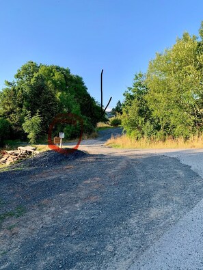 Un chemin de gravier menant à une porte en bois, sur laquelle est apposée une pancarte, le tout étant entouré d’arbres et de végétation.