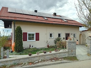 Une maison avec un toit recouvert de tuiles rouges, des panneaux solaires, un petit jardin avec des plantes en pot et un mur en pierre.