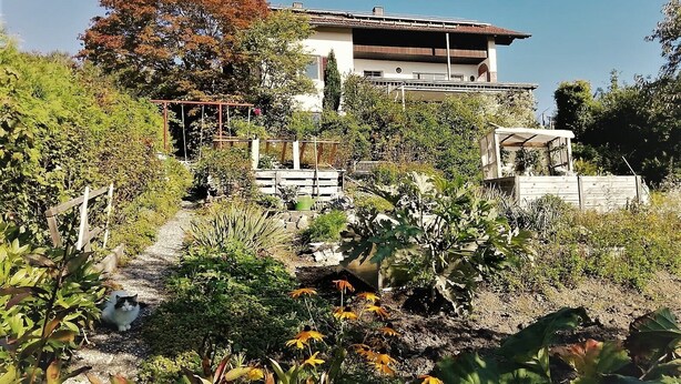 A two-story building with a garden in front, a wooden fence, and a clear sky. - Apartment 'Familie Graml' with Stunning Views, Large Garden with BBQ and Swing (Hauzenberg)
