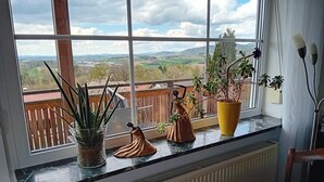 A window with a view of a landscape, a wooden railing, and a table with a candle and a green bowl. - Apartment 'Familie Graml' with Stunning Views, Large Garden with BBQ and Swing (Hauzenberg)