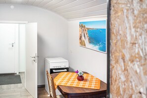 A room with a white door, a white stove, a wooden table with a plaid tablecloth, and a painting of a coastal landscape.