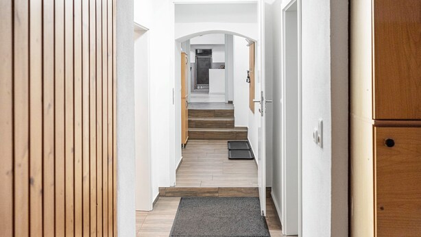 Un pasillo con suelo de madera, un felpudo gris en la entrada y una cocina al fondo.