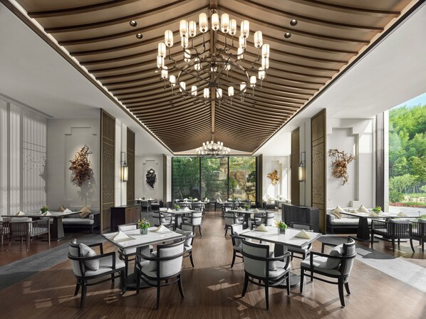 A spacious dining area with multiple tables and chairs, a modern chandelier, and large windows offering a view of greenery outside.
