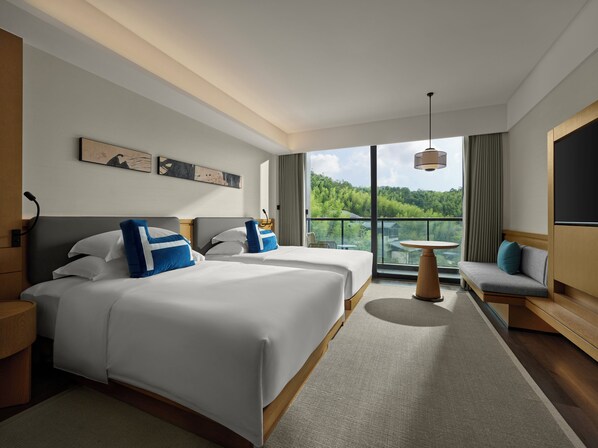 A modern hotel room with two beds, a balcony with a view of a forest, a small round table, and a sofa.