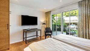 A hotel room with a bed, a desk with a TV, a chair, and a large window with a view of the outdoors. - Apartment with Terrace, Shared Garden, and Wi-Fi (Schmallenberg)