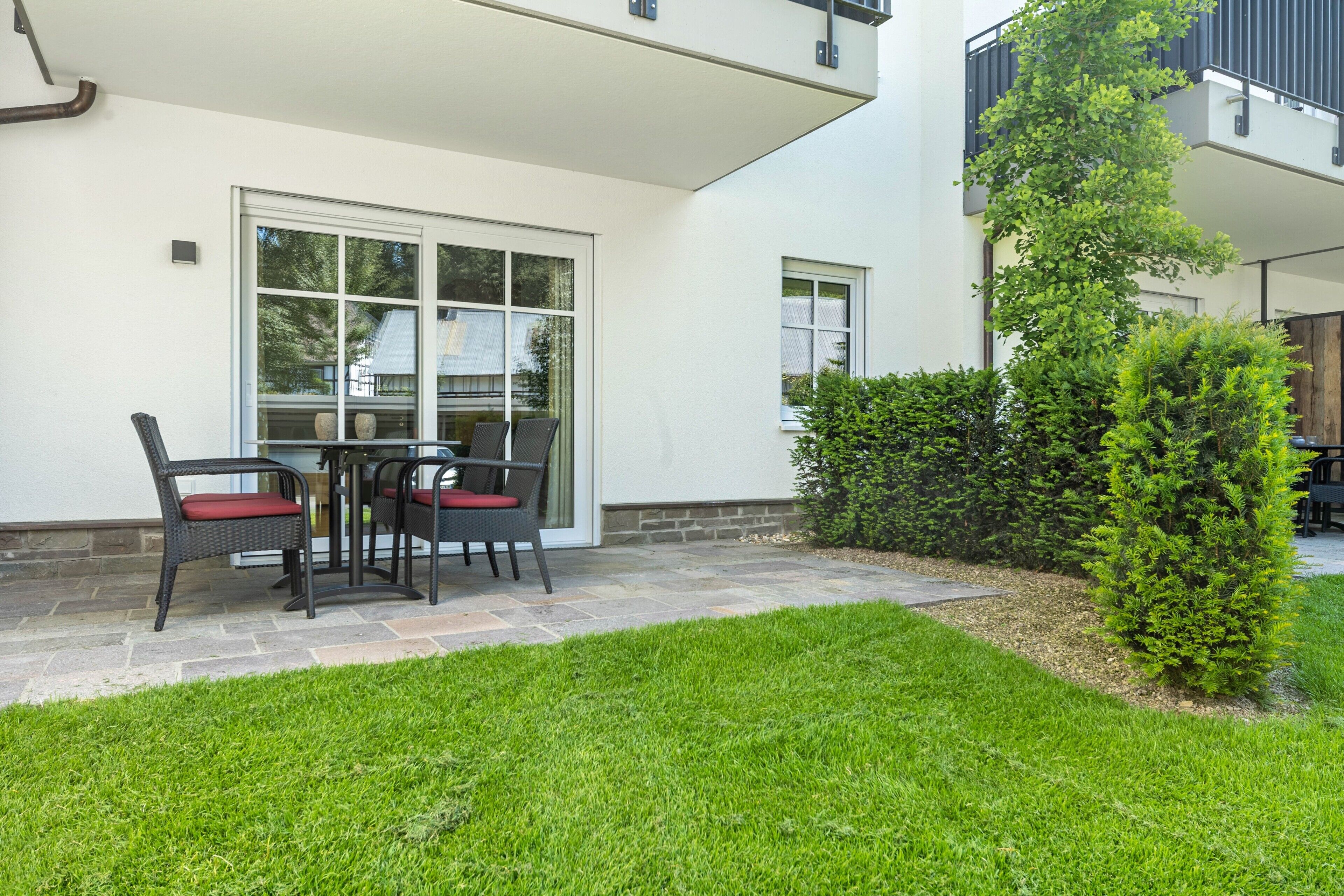 Eine Terrasse mit Tisch und Stühlen, ein kleiner Heckenstreifen und ein Gebäude mit weißer Fassade und einem Fenster.