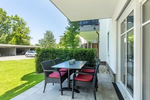 Un patio al aire libre con mesa y sillas, una zona de estar cubierta y vistas a un edificio y vegetación.