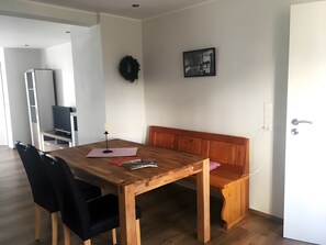 En spiseplads med et træbord, sorte stole, en bænk, et fjernsyn og et billede på væggen.