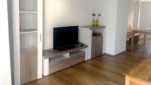 Ein modernes Wohnzimmer mit einem Flachbildfernseher auf einem Holzständer, einem weißen Schrank mit Regalen und einem kleinen Tisch mit zwei Vasen.