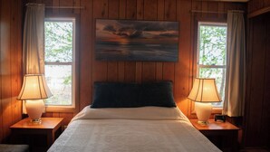 Ein Schlafzimmer mit einem Bett, zwei Nachttischlampen, einem Sonnenuntergangsgemälde und zwei mit Vorhängen versehenen Fenstern.