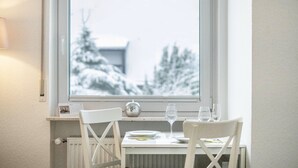 A dining table with chairs, a window with a view of snow-covered trees, and a radiator below the window. - Studio 'Schwarzwaldblick F with Pool App 3' with Balcony and Wi-Fi (Schönwald im Schwarzwald)