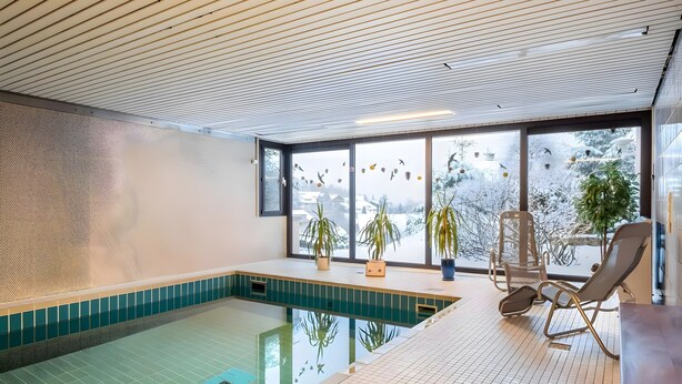 An indoor swimming pool with a tiled floor, a wooden ceiling, and large windows with a view of snow-covered trees. - Studio 'Schwarzwaldblick F with Pool App 3' with Balcony and Wi-Fi (Schönwald im Schwarzwald)