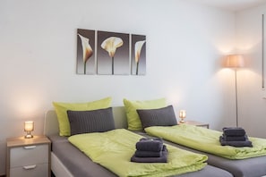 Habitación de hotel con dos camas, cada una con una manta verde y una toalla doblada. Hay dos lámparas de noche, un cuadro floral en la pared y una lámpara de pie alta.