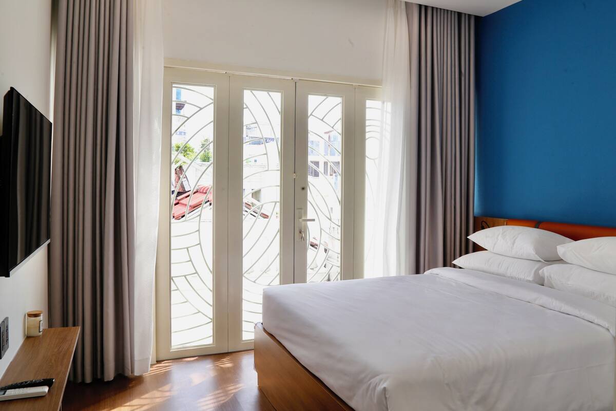 A hotel room with a bed, a TV, a wooden nightstand, and a view through a glass door.