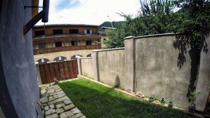 A courtyard with a stone pathway leading to a grassy area, surrounded by a high concrete wall and a wooden building with balconies.