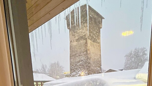 A view through a window with icicles hanging from the ceiling, a stone tower, and snow-covered roofs.