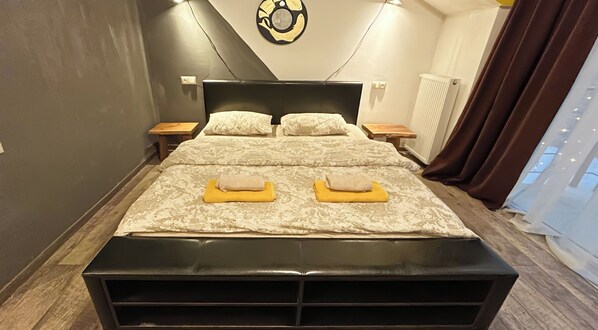 A double bed with a black headboard and footboard, two bedside tables, a wall clock, a radiator, and a window with curtains.