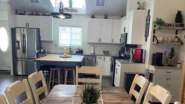 A modern kitchen with white cabinets, a stainless steel refrigerator, a dining table with chairs, and a central island with a plant.
