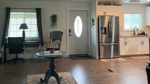 A spacious living room with a ceiling fan, a dining area with a table and chairs, a kitchen with stainless steel appliances, and a door with a frosted glass window.