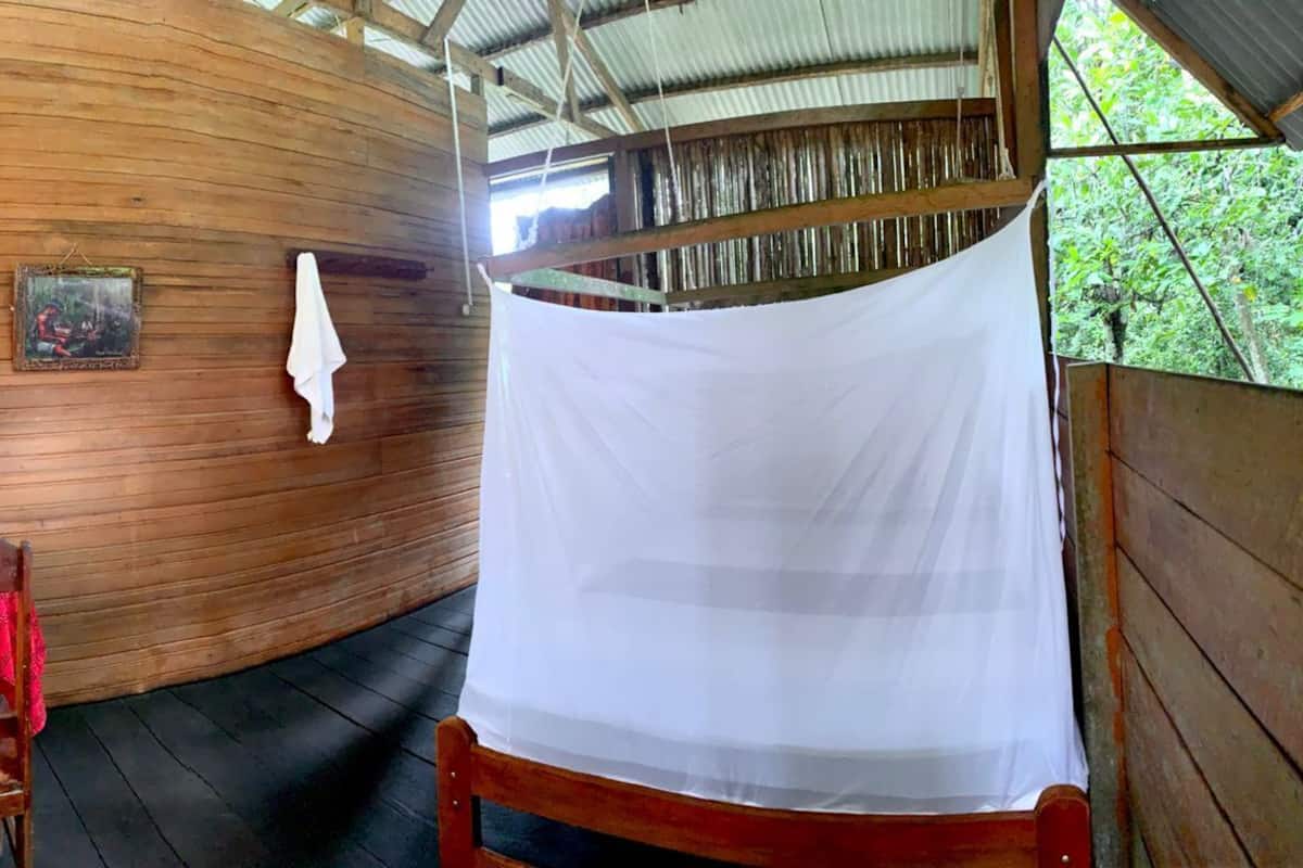 Una cabaña de madera con una pared curva, una pequeña mesa con mantel rojo, una silla, un paño blanco colgado y un cuadro enmarcado.