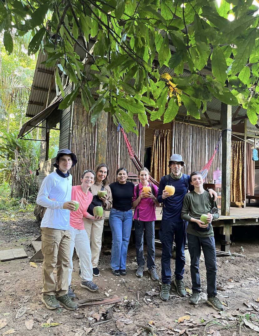 A group of people standing in front of a rustic wooden structure with a thatched roof, surrounded by trees.