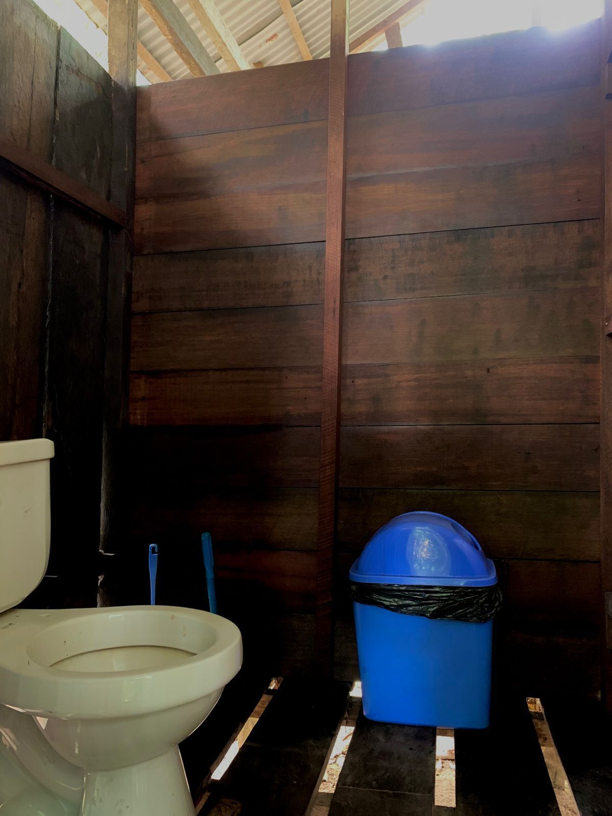 A small, wooden-walled bathroom with a toilet and a blue trash bin.