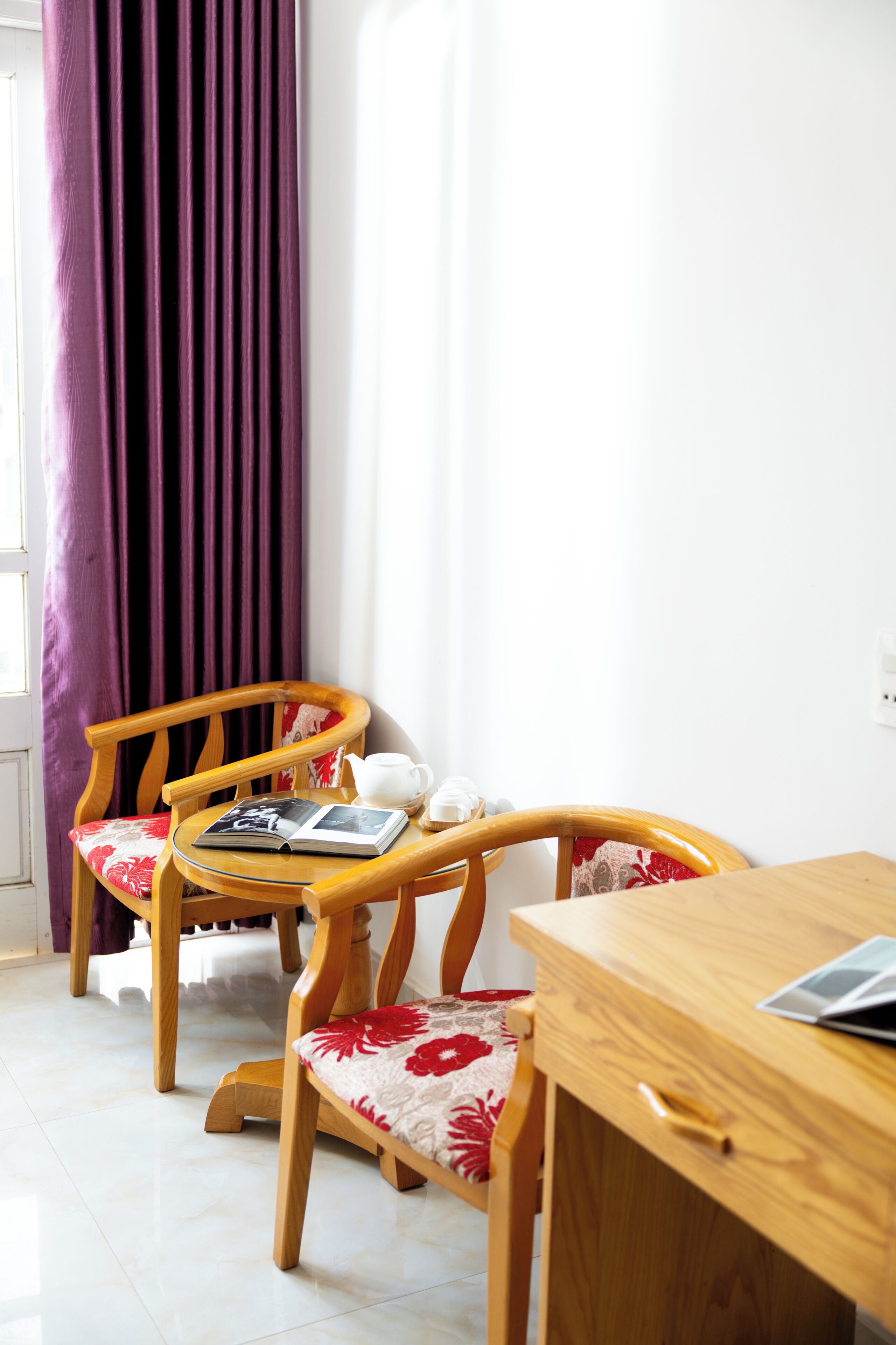 A room with two wooden chairs with floral cushions, a small wooden table with a book and a teapot, and a window with purple curtains.