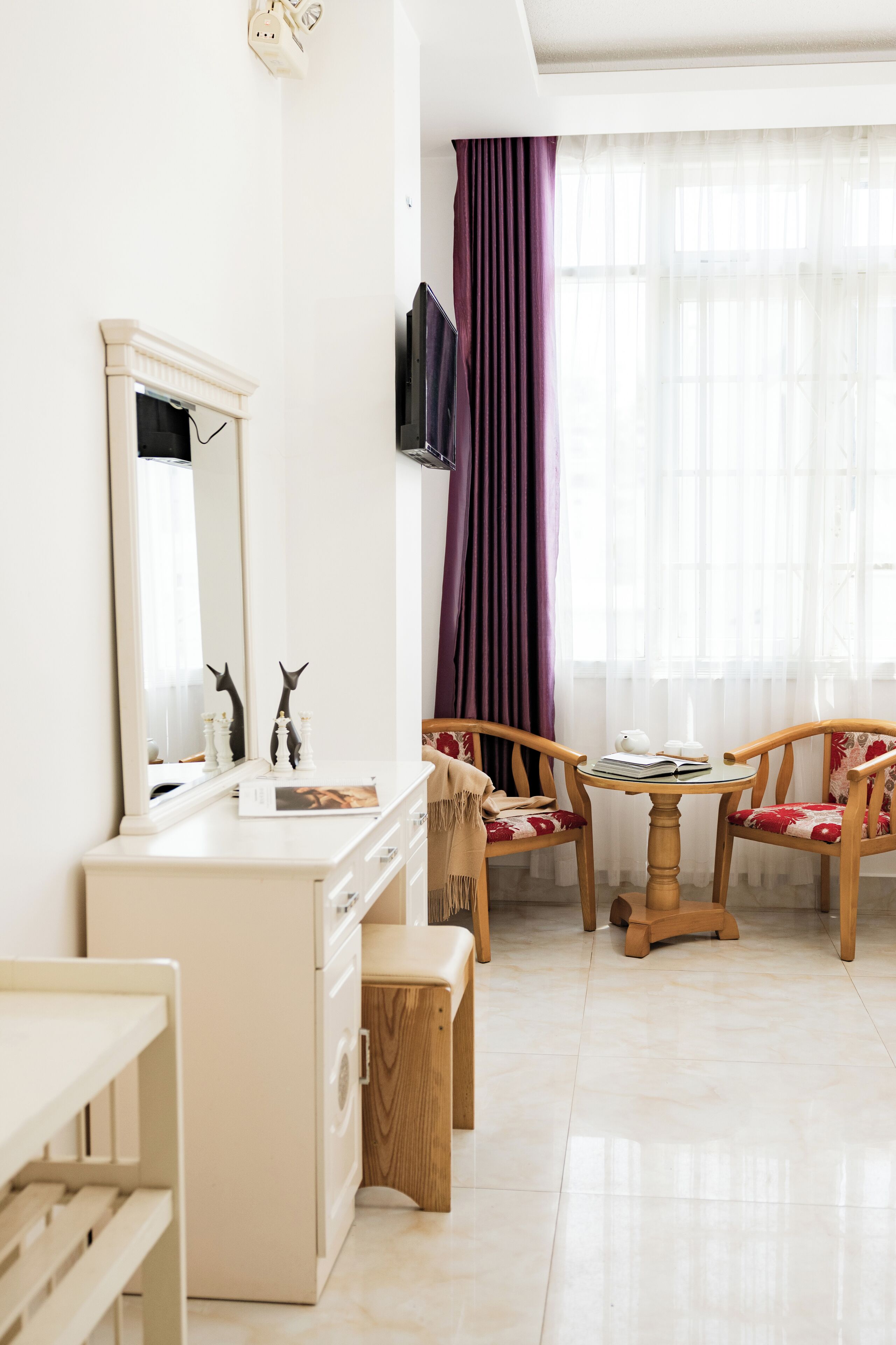 A room with a white vanity, a mirror, a wooden chair with a red cushion, a small round table with a book and a teapot, and a window with purple curtains.