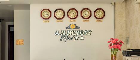 Un mostrador de recepción con relojes que marcan la hora en diferentes ciudades, un letrero que dice "A.M. MEMORY Hotel" y una zona de recepción con un armario de madera y un pequeño santuario.