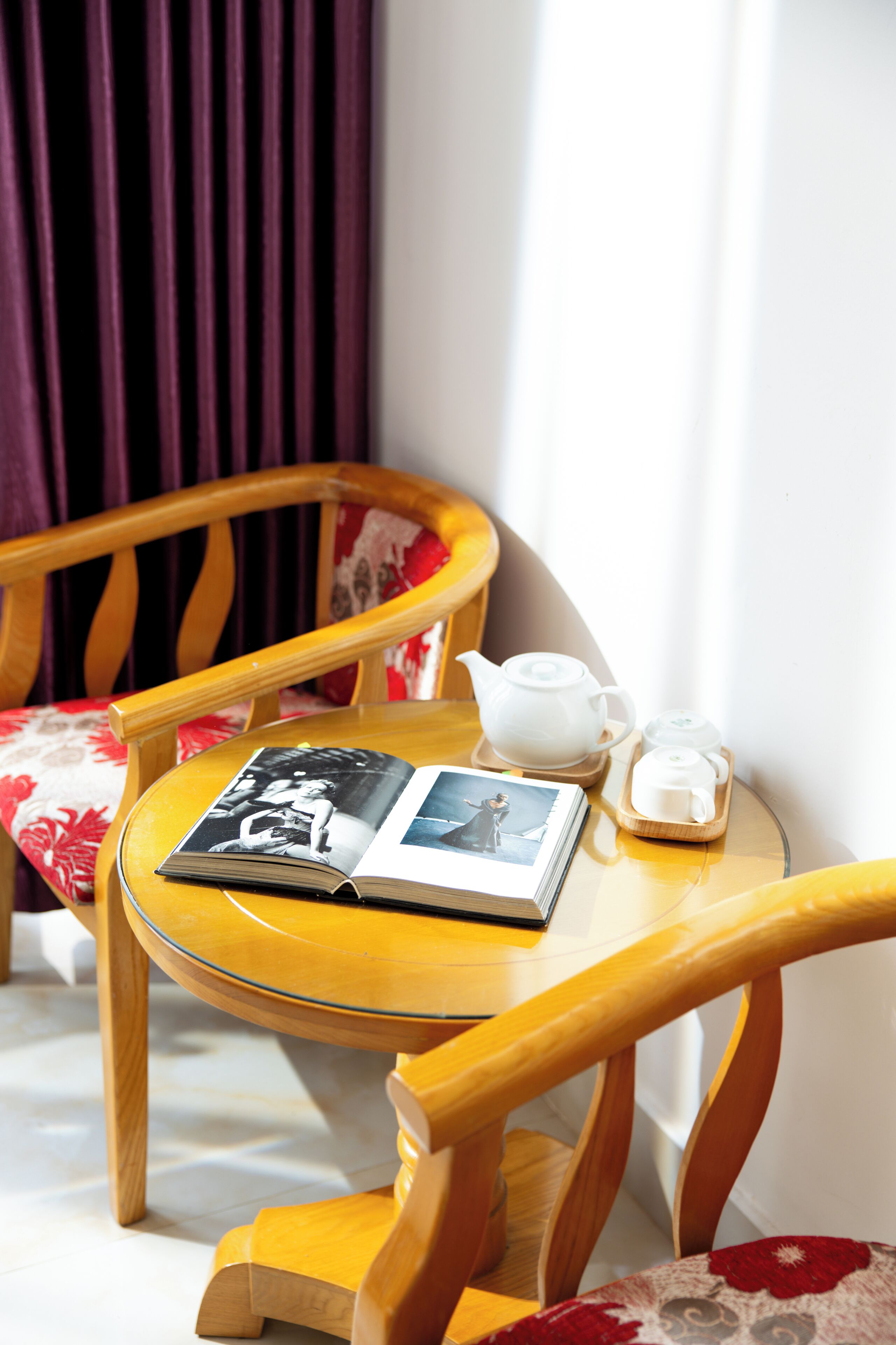Una silla de madera con un cojín floral, una mesita redonda con un libro, una tetera y una taza de té.