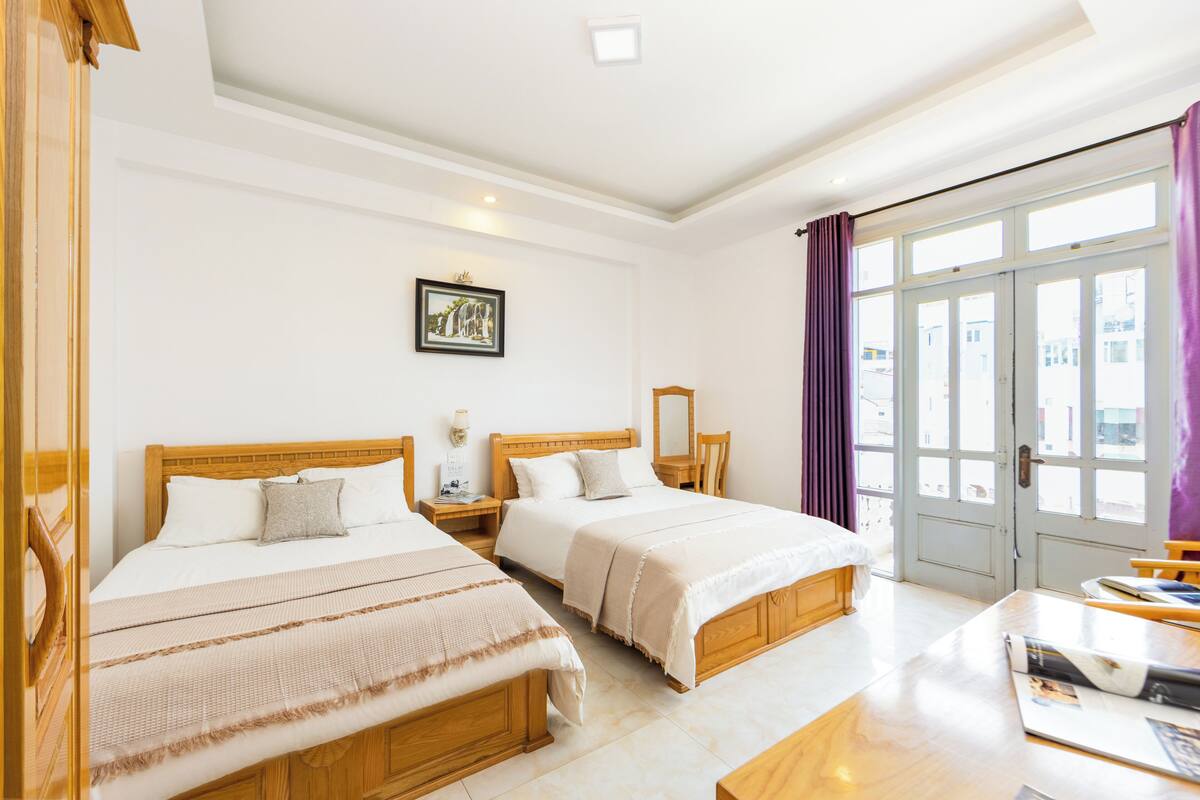 Habitación de hotel con dos camas de madera, un ventanal con cortinas, un escritorio de madera y un cuadro en la pared.