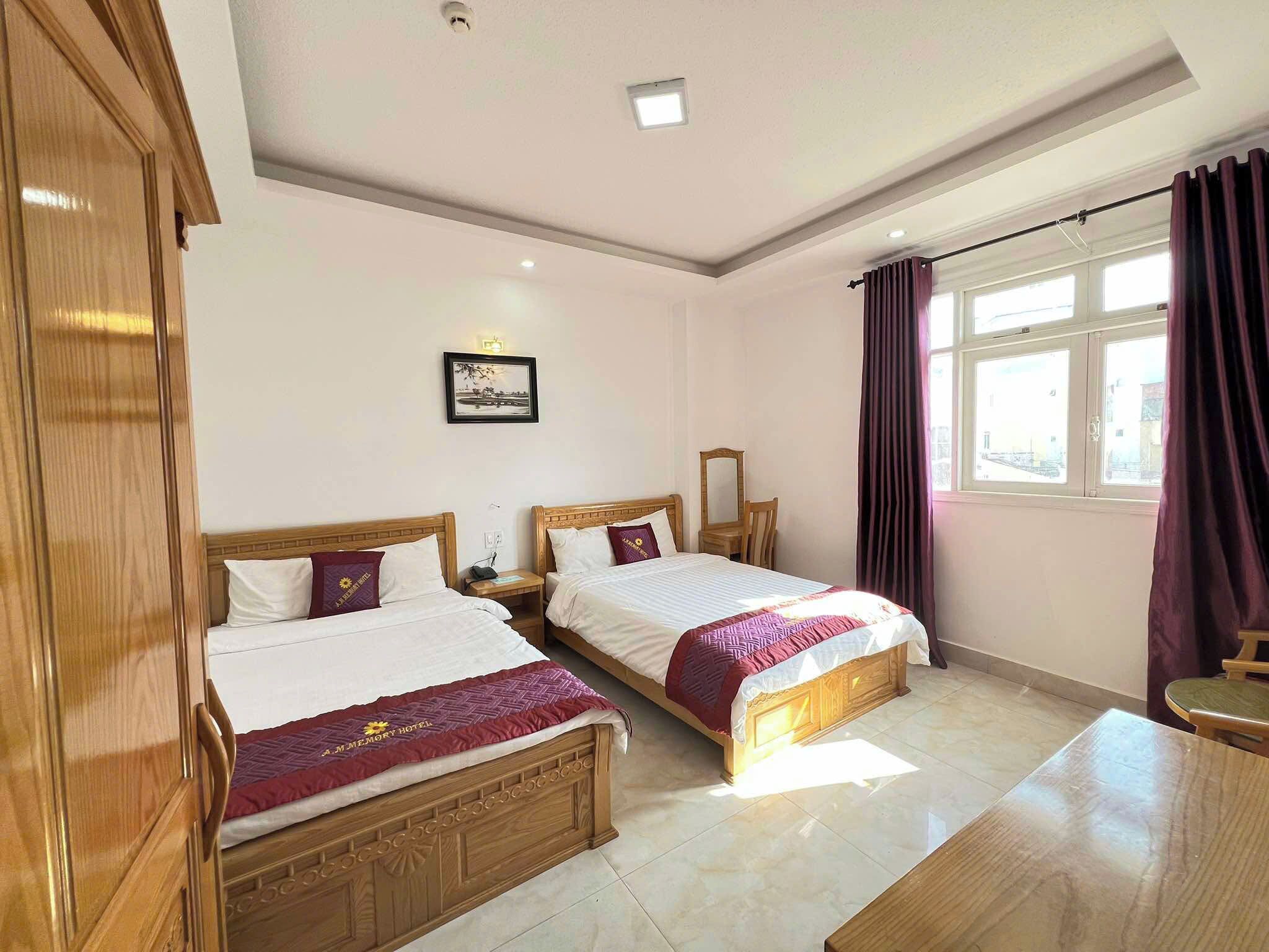 Quarto de hotel com duas camas de madeira, uma mesa pequena, uma cadeira, janela com cortinas e um quadro na parede.