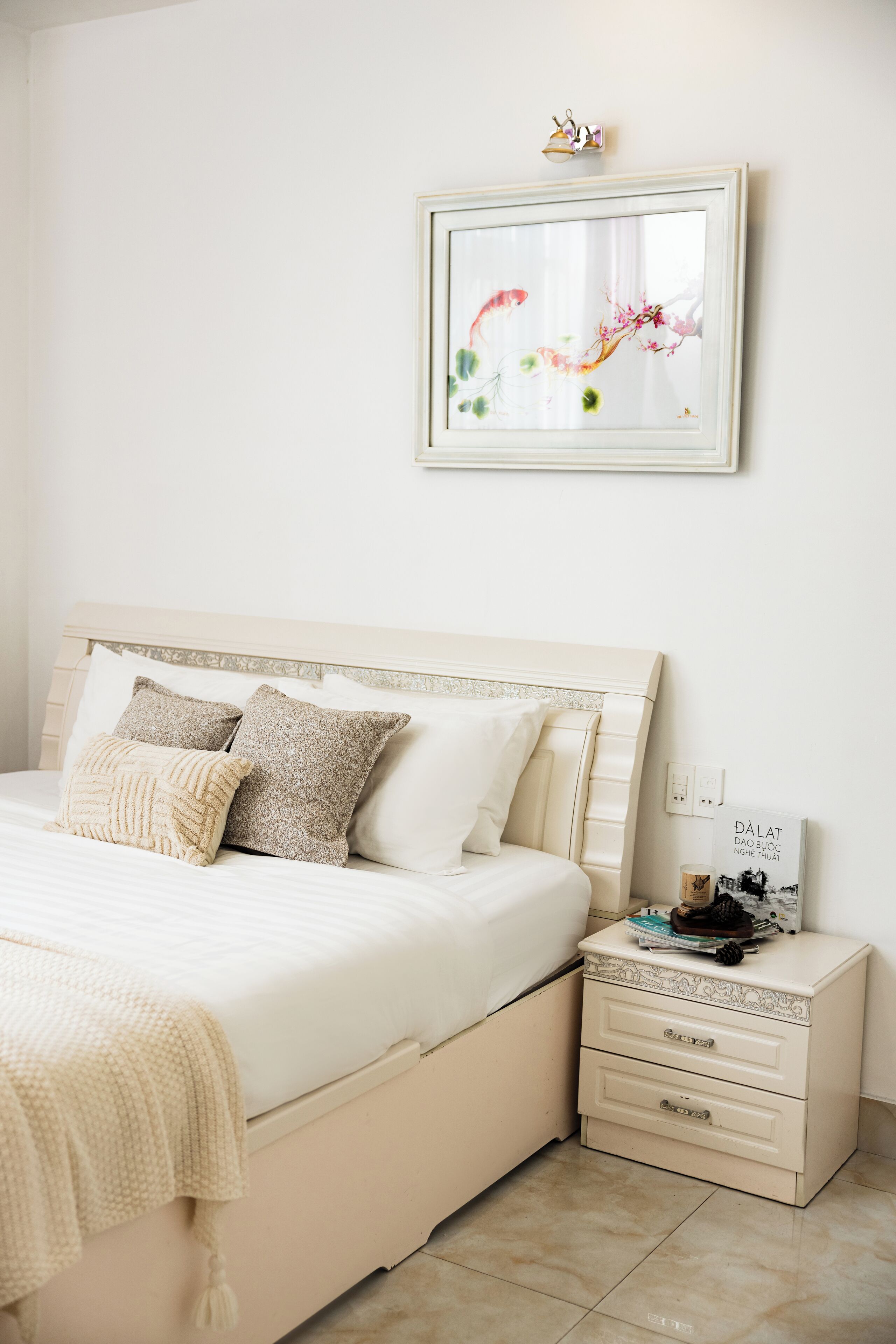 Un dormitorio con cama, almohadas, una mesita de noche con un libro y un cuadro en la pared.