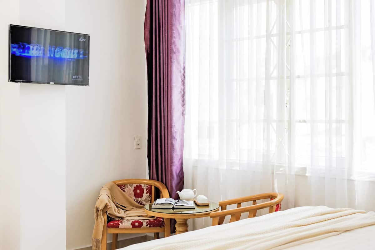 Una habitación de hotel con una cama grande, un televisor montado en la pared, una mesita con una silla y una ventana con cortinas translúcidas.