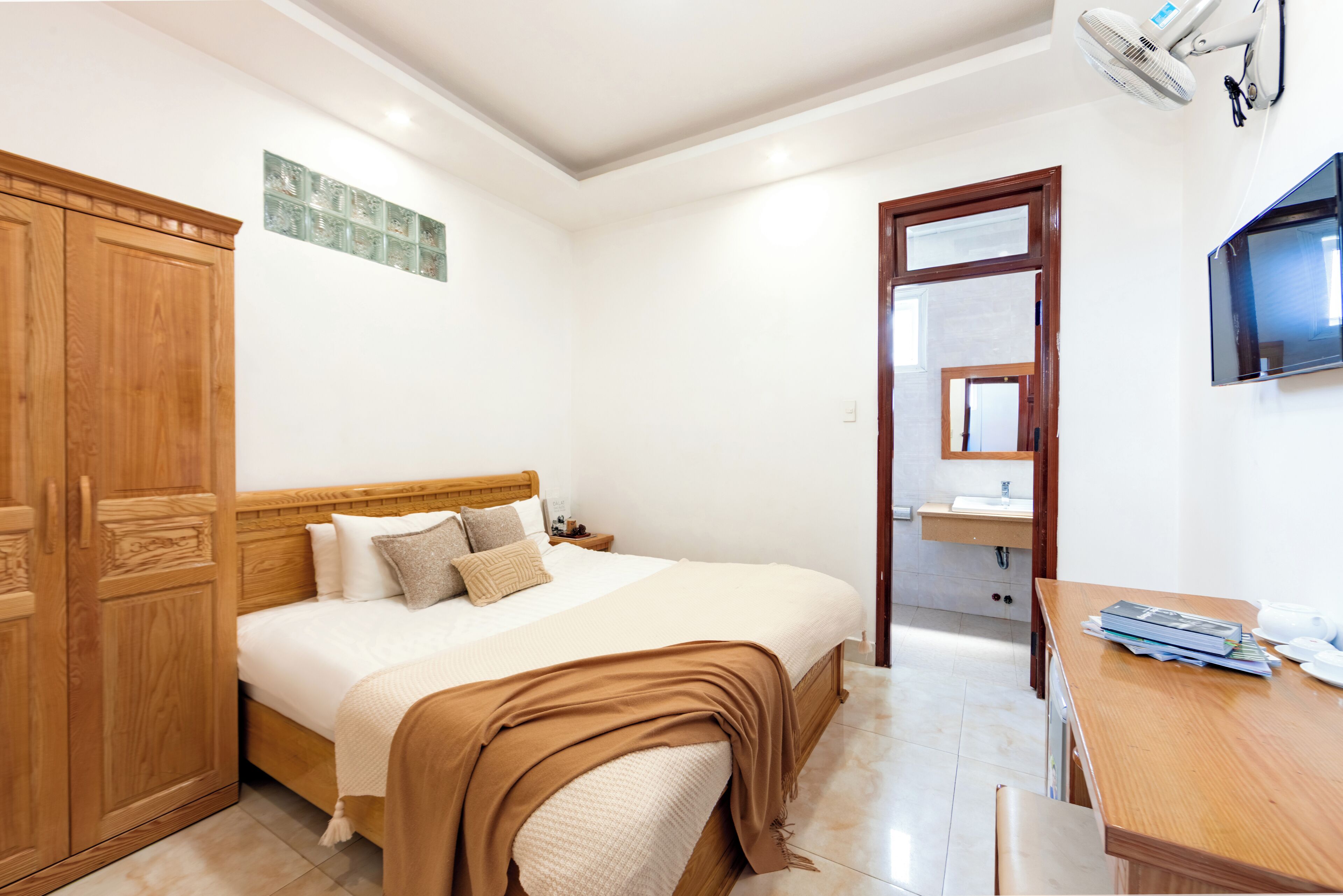 A bedroom with a wooden bed, a wooden wardrobe, a bedside table, a TV, and a bathroom visible through an open door.