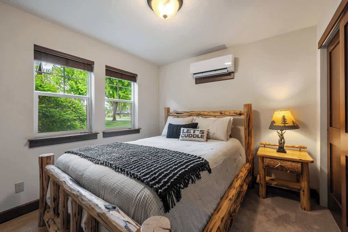 Quarto com cama grande, duas janelas, mesa de cabeceira com luminária e ar-condicionado.
