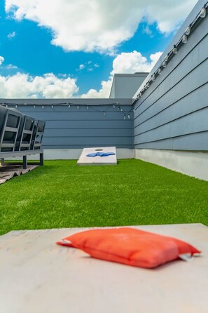 A rooftop terrace with a green lawn, a white table with a red cushion, and a blue pillow on a white mat.