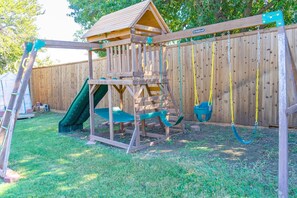 A wooden play structure with a slide and swings, set against a wooden fence.