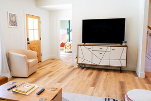 A living room with a sofa, a coffee table with books and remotes, a TV on a cabinet, and a wooden floor. - 3BR Saint Jo Getaway | BBQ, Fire Pit + Comfort (Saint Jo)