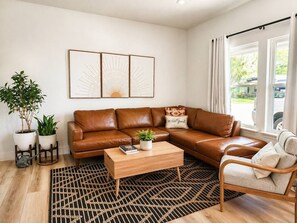 A living room with a brown leather sofa, a wooden coffee table, and a patterned rug. There are two potted plants and a framed artwork on the wall. - 3BR Saint Jo Getaway | BBQ, Fire Pit + Comfort (Saint Jo)