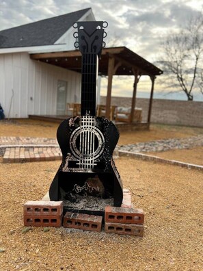 A guitar with a unique design, placed on a gravel surface with bricks around it, in front of a building with a porch. - 3BR Saint Jo Getaway | BBQ, Fire Pit + Comfort (Saint Jo)