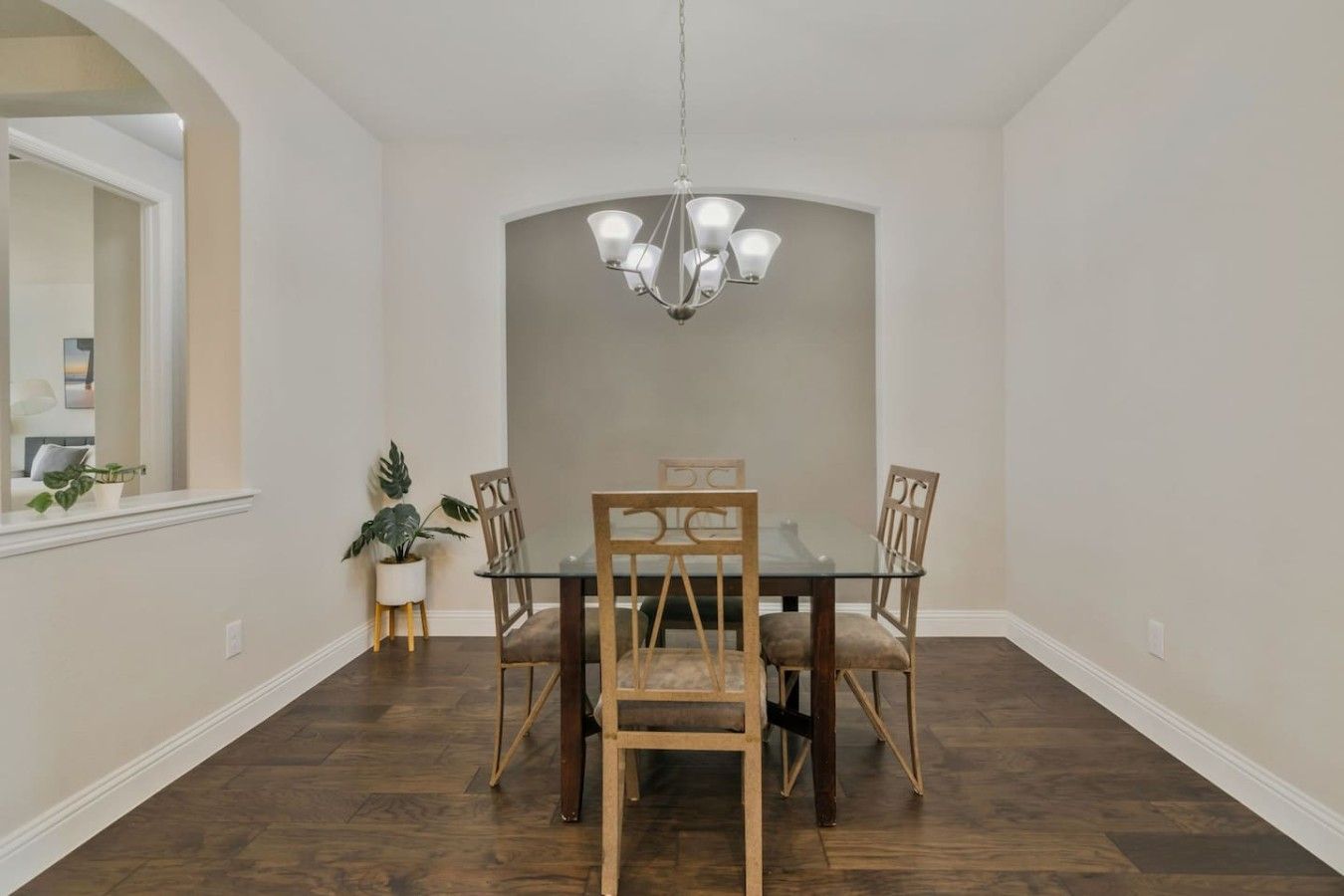 Uma sala de jantar com mesa de vidro, quatro cadeiras de madeira, um lustre e um vaso de planta.