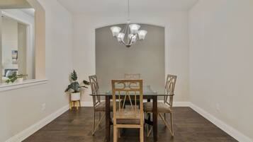 Uma sala de jantar com mesa de vidro, quatro cadeiras de madeira, um lustre e um vaso de planta.