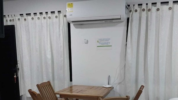 A small room with a wooden table and four chairs, a white bed, and an air conditioning unit on the wall.