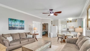 A living room with a beige sofa, a coffee table, and a painting of a beach scene. There is a ceiling fan, a dining table with chairs, and a kitchen area with cabinets and a refrigerator. - Charming 3 BR, 2 BA cottage with pool, perfectly nestled in Orange Beach. (Orange Beach)