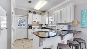 A modern kitchen with white cabinets, a central island with stools, and a decorative wall with motivational phrases. - Charming 3 BR, 2 BA cottage with pool, perfectly nestled in Orange Beach. (Orange Beach)