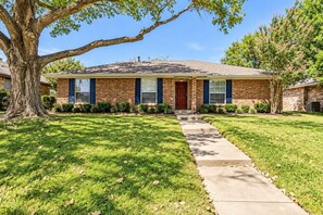 A two-story brick house with a front porch, a large tree, and a well-maintained lawn.