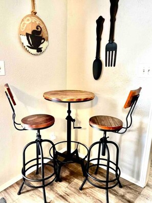 A corner with a small round table and two stools, a wall-mounted coffee-themed sign, and a spatula hanging on the wall.