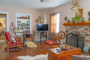 A cozy living room with a fireplace, a television, and a bookshelf filled with various items. - Saint Jo 2BR Wine Country Cottage | Scenic Patio & Cozy Retreat (Saint Jo)